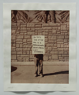 Artist holds sign "On this side of the wall is a brown working class neighborhood" Sign hides upper body,In front of highwall palm trees beyond.