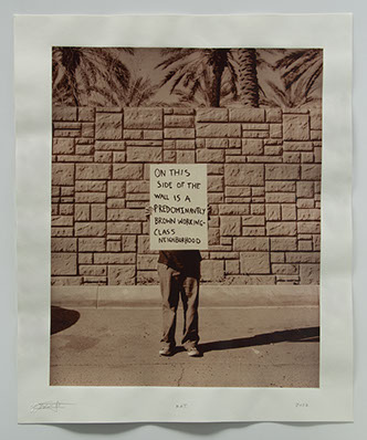 Artist holds sign "On this side of the wall is a brown working class neighborhood" Sign hides upper body,In front of highwall palm trees beyond.