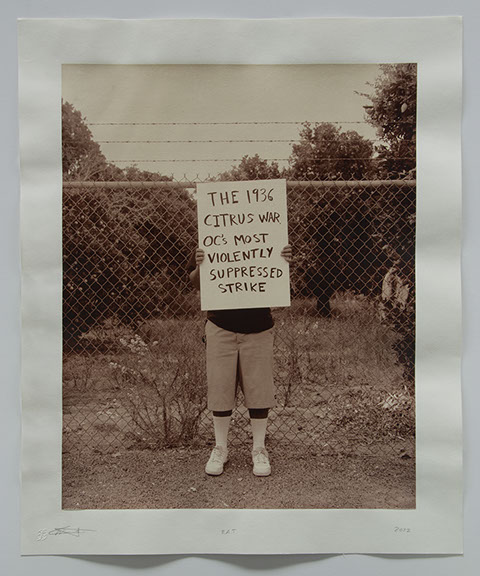 Artist holds sign in front of chainlink fence with citrus trees beyond. Sign says "The 1936 Citrus was OC's most violently suppressed strike"