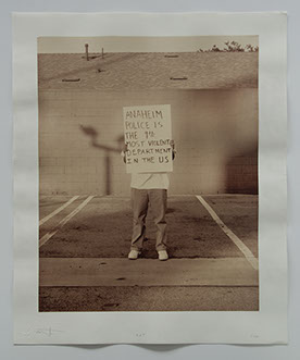 Artist stands in parking lot w concrete wall & roof behind. Obscured by sign he holds: "Anaheim Police is the 9th most violent dept in the US".