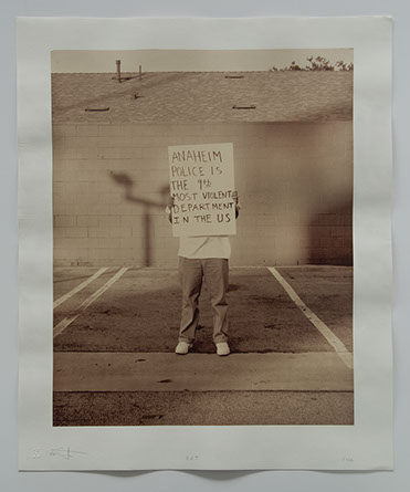 Artist stands in parking lot w concrete wall & roof behind. Obscured by sign he holds: "Anaheim Police is the 9th most violent dept in the US".