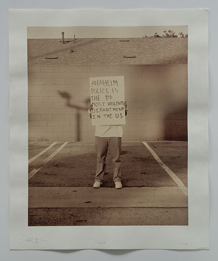 Artist stands in parking lot w concrete wall & roof behind. Obscured by sign he holds: "Anaheim Police is the 9th most violent dept in the US".