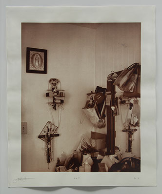 Corner of room with vanity wedged in. Religious objects & mementos cover surface and wall & reflected in mirror.