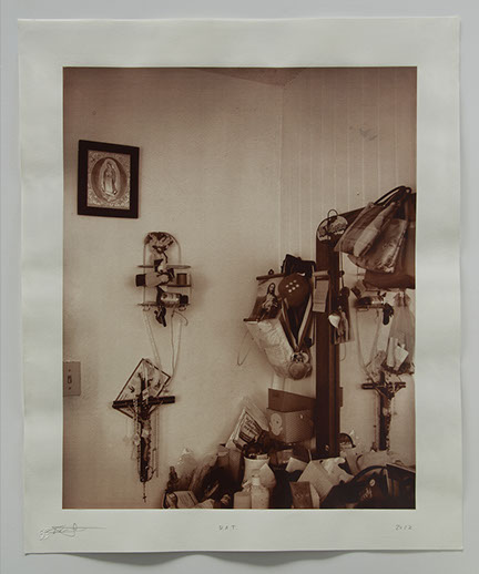 Corner of room with vanity wedged in. Religious objects & mementos cover surface and wall & reflected in mirror.