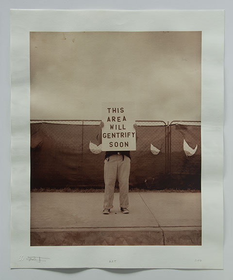 Artist stands w sign on sidewalk in front of chainlink fence covered in plastic. Empty space behind " this area will gentrify soon"