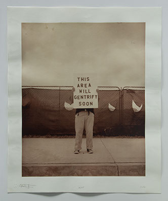 Artist stands w sign on sidewalk in front of chainlink fence covered in plastic. Empty space behind " this area will gentrify soon"