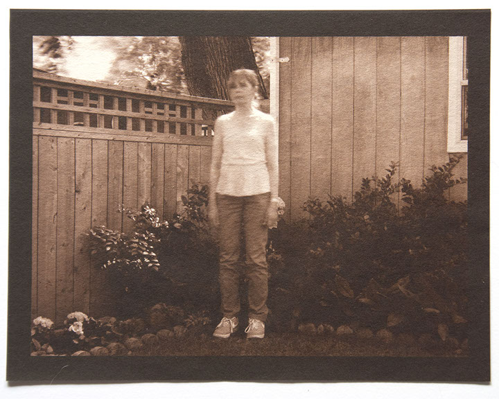 Artist stands center 3/4 view with arms down in corner of backyard. Wood fence, house and plants behind her. All in focus except her upper body 