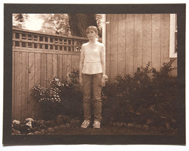 Artist stands center 3/4 view with arms down in corner of backyard. Wood fence, house and plants behind her. All in focus except her upper body 
