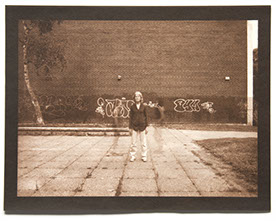 Artist stands ctr facing forward. Large brick wall with graffiti behind. Square pavers with weeds form a grid below. 2 ghost figures flank her.