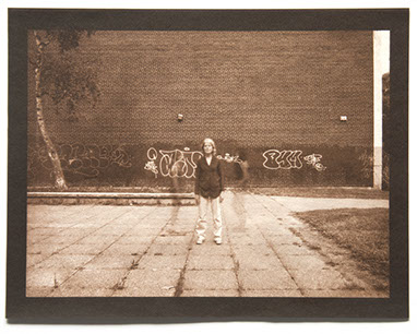 Artist stands ctr facing forward. Large brick wall with graffiti behind. Square pavers with weeds form a grid below. 2 ghost figures flank her.