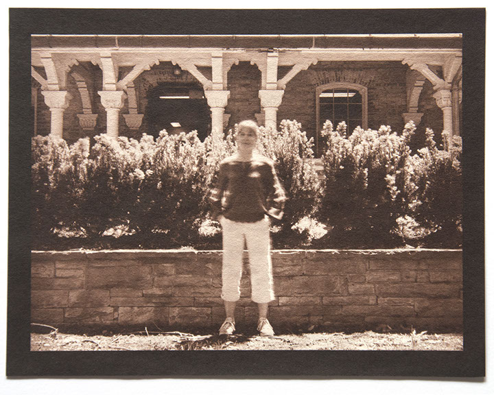Artist stands ctr face forward in bright day. Behind her a low wall /w small shrubs reaches her height. Backgrnd is an ornate wood & stone porch
