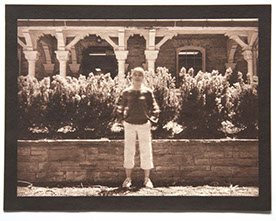 Artist stands ctr face forward in bright day. Behind her a low wall /w small shrubs reaches her height. Backgrnd is an ornate wood & stone porch