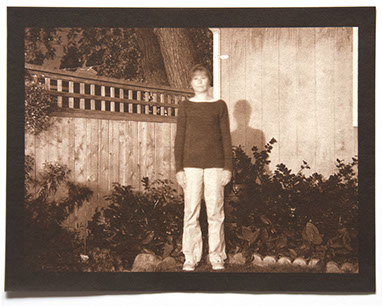 Artist stands in same position in backyard surround by wood fence, house, shrubs. It is nighttime. Brightly lit /w her shadow on wall behind her