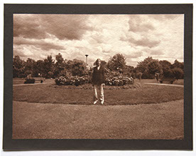 Artist stands ctr facing forward in round landscape of grass. Flowering shrubs at center behind her. Rolling clouds above and trees in distance.