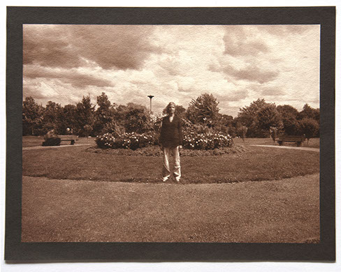 Artist stands ctr facing forward in round landscape of grass. Flowering shrubs at center behind her. Rolling clouds above and trees in distance.