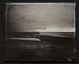 Panel C2-Land, grasses & shoreline dominate bottom half. Houses near the shore. Sky heavy with clouds with 1 clear sliver over ocean in distance