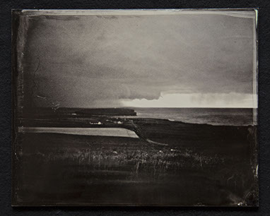 Panel C2-Land, grasses & shoreline dominate bottom half. Houses near the shore. Sky heavy with clouds with 1 clear sliver over ocean in distance