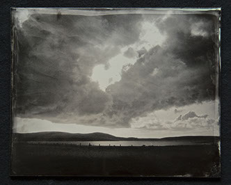 Panel C3 - Big sky full of dark clouds. Light peaks through and below. A low horizon of landscape with a lake surrounded by land and hills.