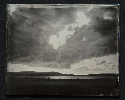 Panel C3 - Big sky full of dark clouds. Light peaks through and below. A low horizon of landscape with a lake surrounded by land and hills.